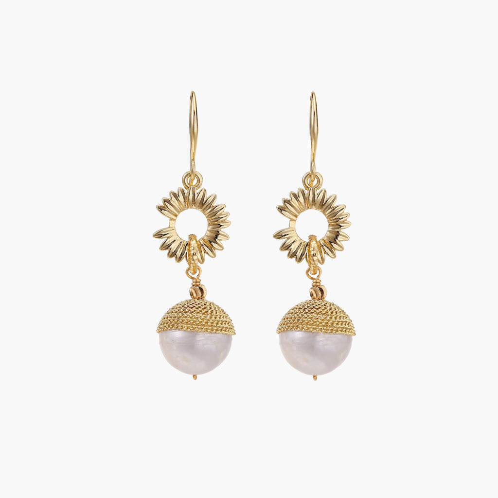 Gold sunburst charm earrings with baroque pearl drops on French hooks against cream background