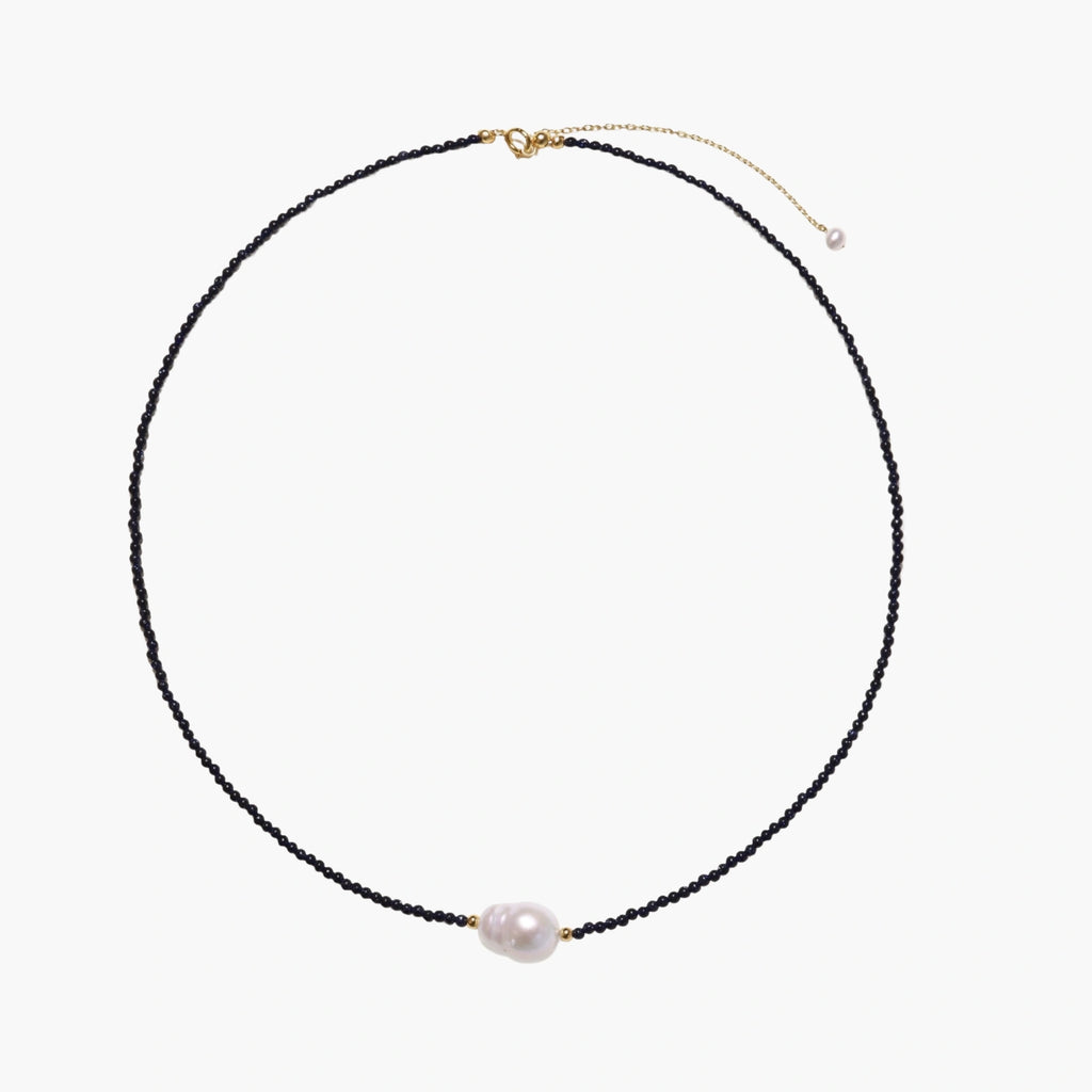 Delicate necklace with black blue sandstone beads and baroque pearl center on white background with gold accents.