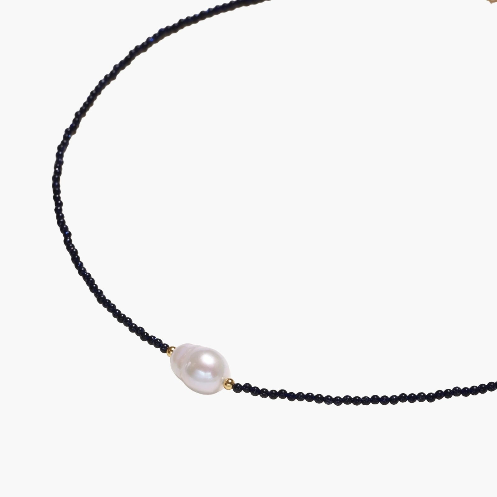 Delicate necklace with black blue sandstone beads and baroque pearl center on white background with gold accents.