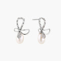 Silver bow-shaped stud earrings with baguette crystals and dangling white freshwater pearls on cream background
